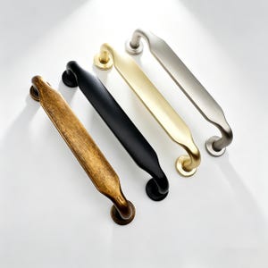 Pode incluir: Quatro puxadores de armário em diferentes acabamentos: latão antigo, preto, dourado e níquel escovado. Cada puxador tem uma alça curva e placas de montagem circulares. Os puxadores estão dispostos diagonalmente em uma superfície branca, mostrando suas cores e designs.