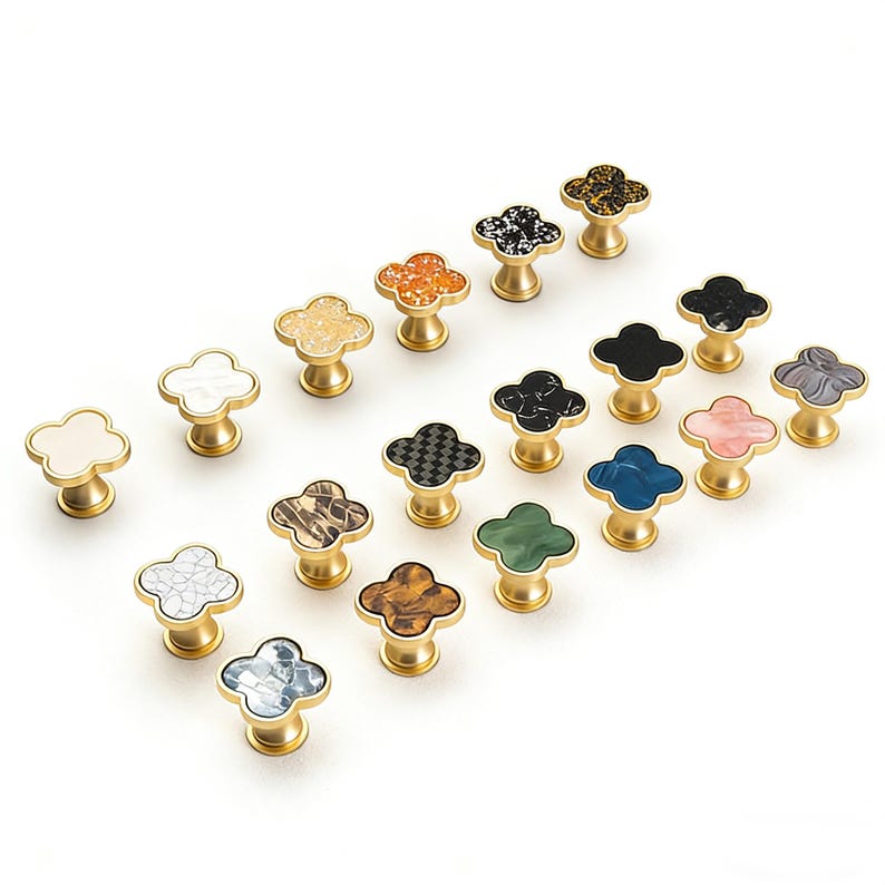 K&ouml;nnte beinhalten: Eine Sammlung dekorativer Schrankkn&ouml;pfe mit einem vierbl&auml;ttrigen Kleeblatt-Design. Die Kn&ouml;pfe weisen verschiedene Farben und Muster auf, darunter Wei&szlig;, Schwarz, Orange und Gr&uuml;n, auf einer goldfarbenen Basis. Diese Kn&ouml;pfe sind ideal, um M&ouml;beln einen Hauch von Eleganz zu verleihen.