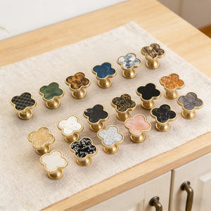 K&ouml;nnte beinhalten: Eine Sammlung dekorativer Schrankkn&ouml;pfe mit Kleeblatt-Design. Die Kn&ouml;pfe weisen verschiedene Farben und Muster auf, darunter Schwarz, Wei&szlig;, Gold und marmor&auml;hnliche Texturen. Jeder Knopf hat einen messingfarbenen Sockel.