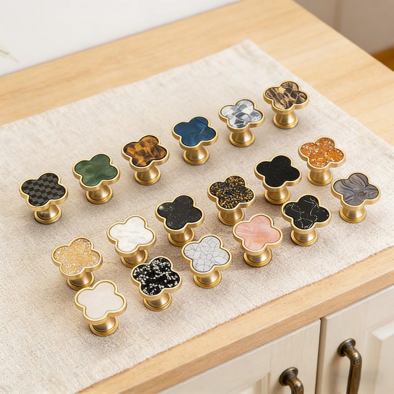 K&ouml;nnte beinhalten: Eine Sammlung dekorativer Schrankkn&ouml;pfe mit Kleeblatt-Design. Die Kn&ouml;pfe weisen verschiedene Farben und Muster auf, darunter Schwarz, Wei&szlig;, Gold und marmor&auml;hnliche Texturen. Jeder Knopf hat einen messingfarbenen Sockel.