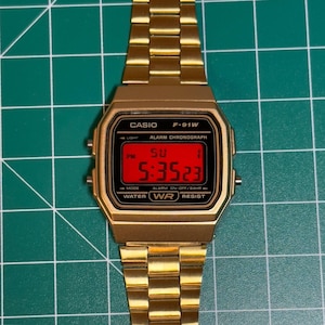 May include: A gold-toned Casio F-91W digital watch with a rectangular red display showing the time. The watch has a gold metal band and a black bezel with white text. The time displayed is 5:35:23 PM on Sunday.