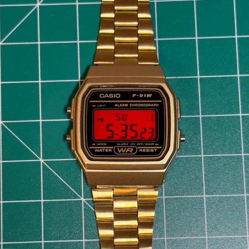 Peut inclure: Montre num&eacute;rique Casio F-91W dor&eacute;e avec un &eacute;cran rouge rectangulaire affichant l'heure. La montre a un bracelet en m&eacute;tal dor&eacute; et un bo&icirc;tier noir avec du texte blanc. L'heure affich&eacute;e est 17h35m23s le dimanche.