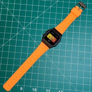 May include: A digital watch with a black face and an orange strap. The watch shows the time as 9:30. The strap is made of a flexible material and has a silver buckle. The watch is on a green surface.