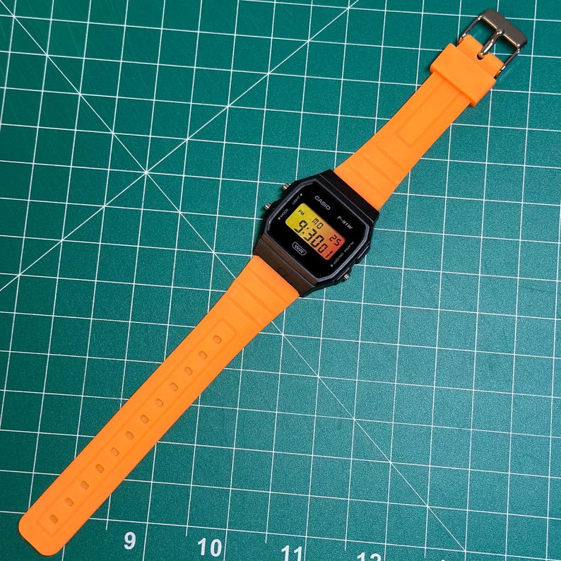 May include: A digital watch with a black face and an orange strap. The watch shows the time as 9:30. The strap is made of a flexible material and has a silver buckle. The watch is on a green surface.