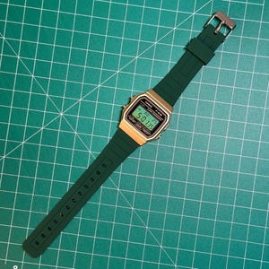 May include: A digital watch with a gold-colored face and a dark green band. The watch displays the time and date on a digital screen. The band is made of a flexible material and has a silver-colored buckle. The watch is set against a green grid background.