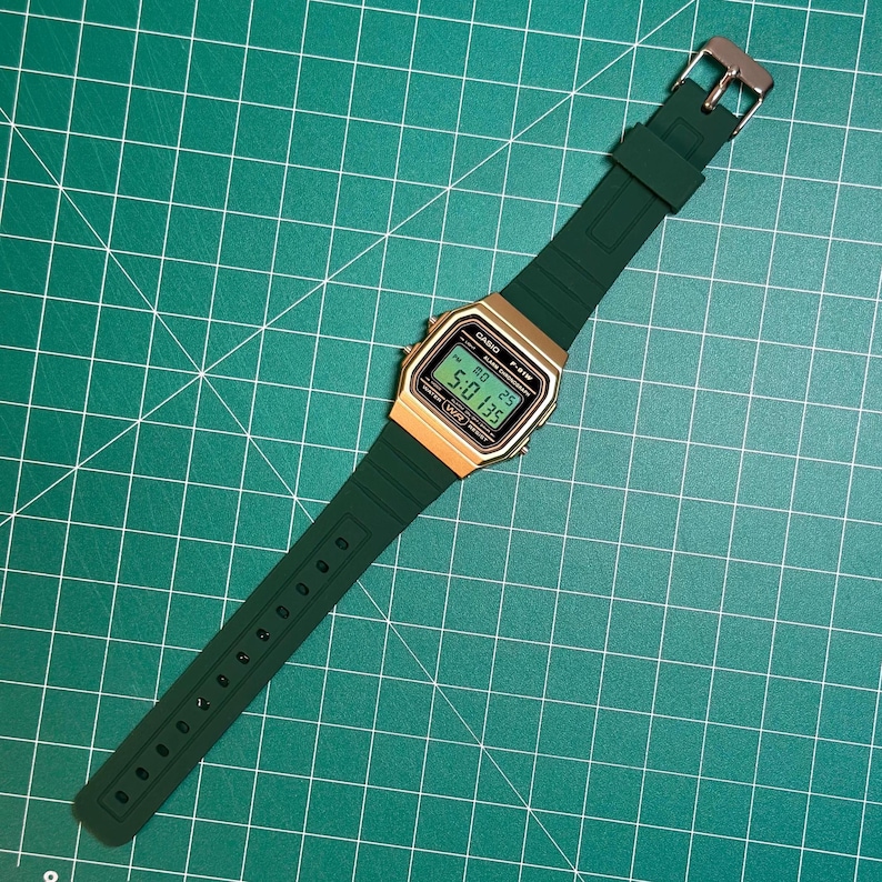 May include: A digital watch with a gold-colored face and a dark green band. The watch displays the time and date on a digital screen. The band is made of a flexible material and has a silver-colored buckle. The watch is set against a green grid background.