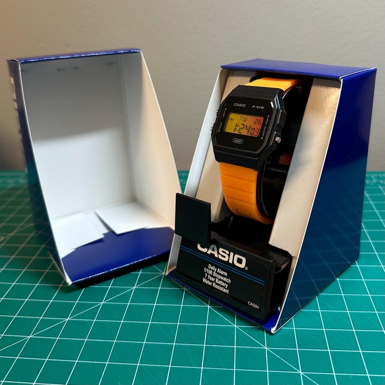May include: A black Casio digital watch with an orange strap is presented in its blue and white box. The watch face displays the time and date. The box is open, showing the watch and the Casio brand details.