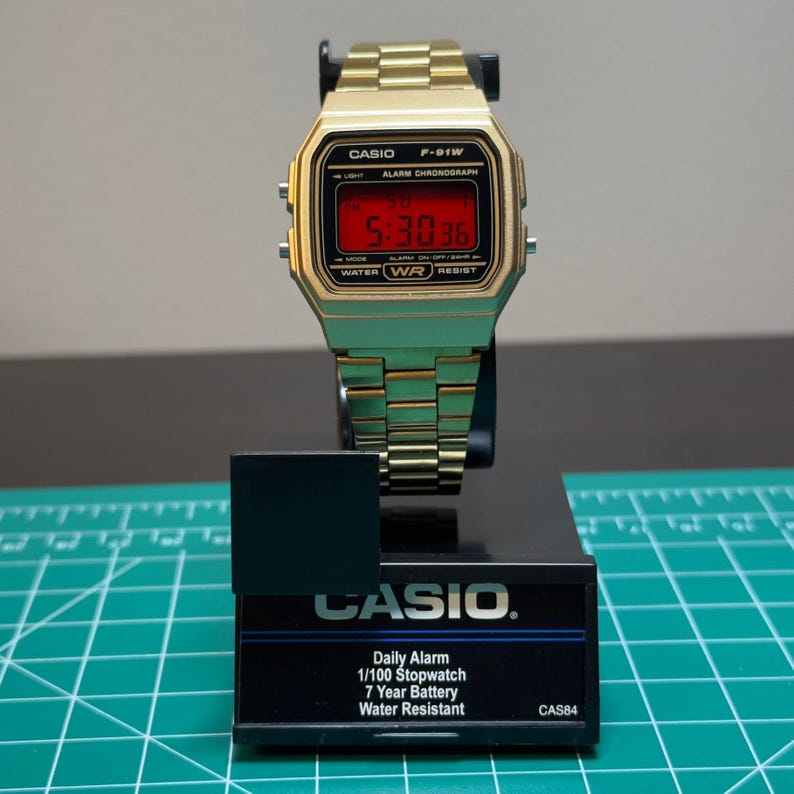 Peut inclure: Montre num&eacute;rique Casio F-91W dor&eacute;e avec un affichage num&eacute;rique rouge indiquant l'heure. La montre a un cadran rectangulaire et un bracelet en m&eacute;tal dor&eacute;. La montre est pr&eacute;sent&eacute;e sur un support noir avec le logo Casio et les caract&eacute;ristiques.
