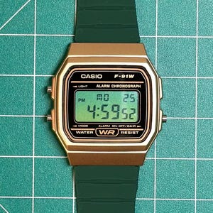 May include: A gold-toned Casio F-91W digital watch with a green strap. The watch face displays the time, day, and date. The watch has the text "CASIO F-91W ALARM CHRONOGRAPH" and "WATER RESIST" printed on the face.