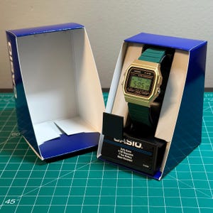 May include: A gold-toned Casio F-91W digital watch with a green strap is displayed in its original blue box. The watch face shows the time as 5:05:18. The box includes the Casio logo and watch specifications.