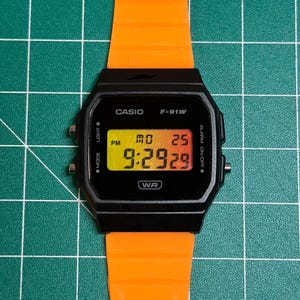 May include: Black Casio F-91W digital watch with an orange band. The watch face displays the time, day, and date. The watch has buttons for mode, light, and alarm functions. The watch is water resistant.