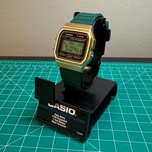 May include: A gold-toned Casio F-91W digital watch with a green band is displayed on a black stand. The watch face shows the time, date, and other functions. The watch has the text "CASIO" and "F-91W" on the face. The stand has the text "CASIO" and watch features.