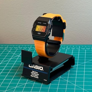 May include: A black Casio digital watch with an orange strap is displayed on a black stand. The watch face shows the time and date. The stand has the Casio logo and product information printed on it.