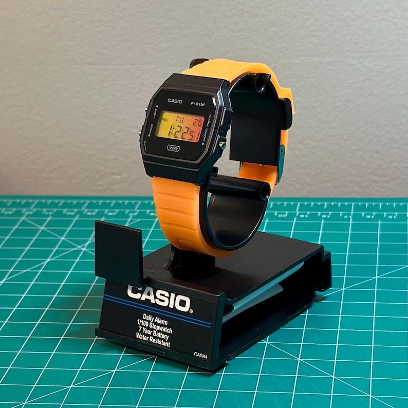 May include: A black Casio digital watch with an orange strap is displayed on a black stand. The watch face shows the time and date. The stand has the Casio logo and product information printed on it.