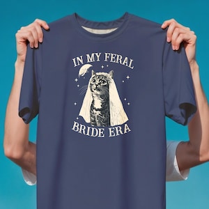 May include: Navy blue t-shirt featuring a cat wearing a veil, with the text "IN MY FERAL BRIDE ERA" in white. The design includes a crescent moon and stars. The shirt is held up against a blue background.