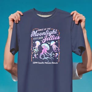 May include: Navy blue t-shirt featuring a graphic of jellyfish in shades of pink, purple, and blue. The text reads "Dance of the Moonlight Jellies, August 28th, 10PM South Pelican Beach."