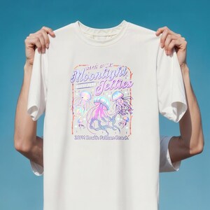 May include: White t-shirt featuring a colorful graphic of jellyfish with the text "Dance of the Moonlight Jellies" and "10PM South Pelican Beach". The design includes pink, purple, and blue hues, creating a vibrant and eye-catching design.