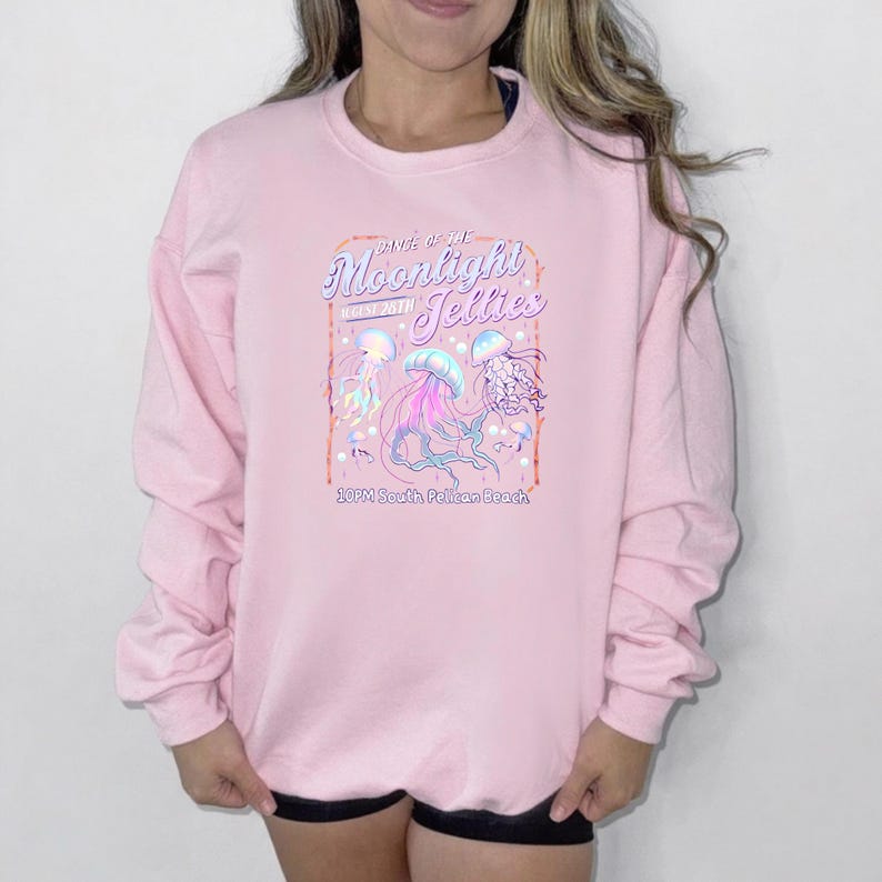 May include: A light pink sweatshirt with a graphic of jellyfish in shades of blue, pink, and purple. The text on the sweatshirt reads "Dance of the Moonlight Jellies" and "10PM South Pelican Beach".