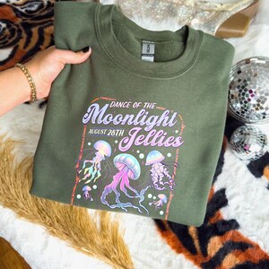 May include: An olive green sweatshirt featuring a colorful graphic of jellyfish with the text "Dance of the Moonlight Jellies August 28th". The design includes pink, purple, and blue jellyfish.