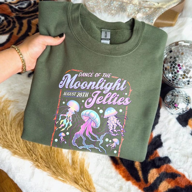 May include: An olive green sweatshirt featuring a colorful graphic of jellyfish with the text "Dance of the Moonlight Jellies August 28th". The design includes pink, purple, and blue jellyfish.