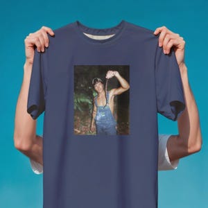 May include: Navy blue t-shirt featuring a printed image of a person holding a snake. The person is wearing denim overalls. The background is a blurred forest scene.