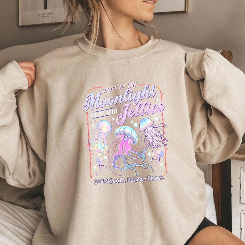 May include: Beige sweatshirt featuring a colorful graphic of jellyfish with the text "Dance of the Moonlight Jellies, August 28th, 10PM South Pelican Beach". The design includes pink, purple, and blue jellyfish.