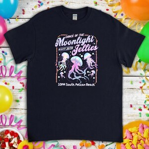 May include: A navy blue t-shirt featuring a graphic design with the text "Dance of the Moonlight Jellies" and the date August 26th. The design includes colorful jellyfish illustrations in shades of pink, blue, and purple. The text "10PM South Pelican Beach" is also included.