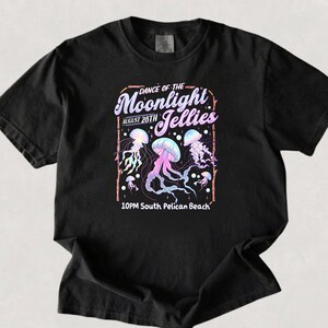 May include: Black t-shirt featuring a graphic design with the text "Dance of the Moonlight Jellies" and "August 28th". The design includes illustrations of jellyfish in shades of purple, blue, and pink. The text "10PM South Pelican Beach" is also included.