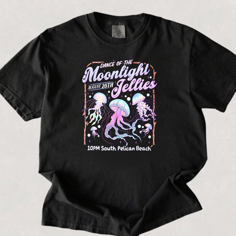 May include: Black t-shirt featuring a graphic design with the text "Dance of the Moonlight Jellies" and "August 28th". The design includes illustrations of jellyfish in shades of purple, blue, and pink. The text "10PM South Pelican Beach" is also included.