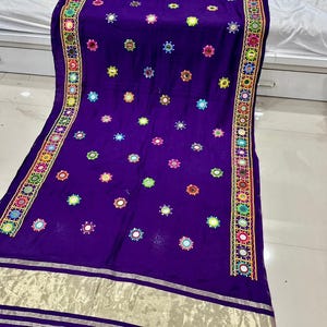 May include: A deep purple textile with detailed embroidery and mirror embellishments. The fabric displays a repeating pattern of colourful circular designs and is bordered with a decorative trim. The edges are finished with a gold-coloured trim.