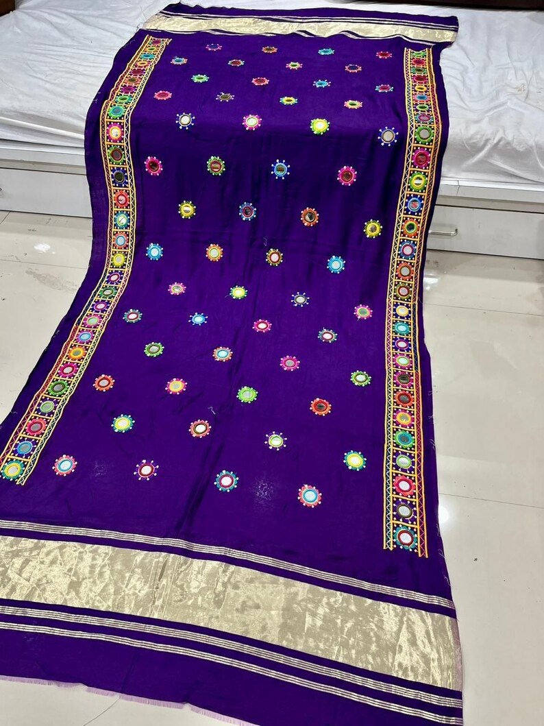 May include: A deep purple textile with detailed embroidery and mirror embellishments. The fabric displays a repeating pattern of colourful circular designs and is bordered with a decorative trim. The edges are finished with a gold-coloured trim.