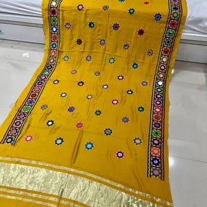 May include: Mustard yellow textile with detailed embroidery and mirror embellishments. The edges have a colourful border with stitching and small mirrors. The fabric is decorated with scattered circular mirror accents, creating a vibrant and decorative design.