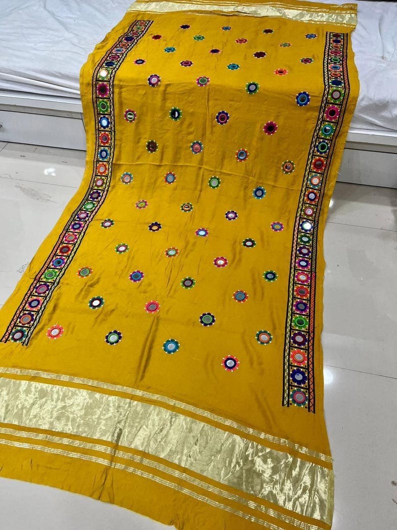 May include: Mustard yellow textile with detailed embroidery and mirror embellishments. The edges have a colourful border with stitching and small mirrors. The fabric is decorated with scattered circular mirror accents, creating a vibrant and decorative design.