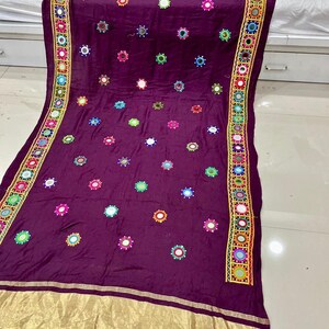 May include: A rich purple textile adorned with numerous circular mirror details. The borders are embellished with a colourful geometric design incorporating small mirrors. A gold band runs along the bottom edge, providing a luxurious finish.