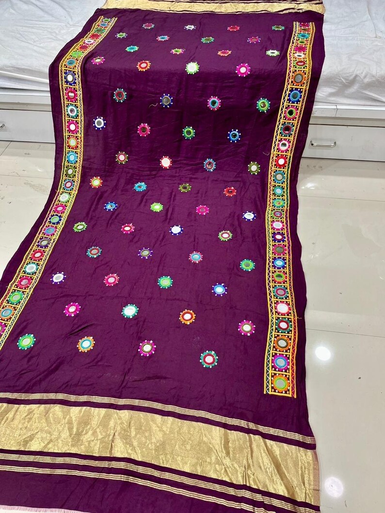 May include: A rich purple textile adorned with numerous circular mirror details. The borders are embellished with a colourful geometric design incorporating small mirrors. A gold band runs along the bottom edge, providing a luxurious finish.