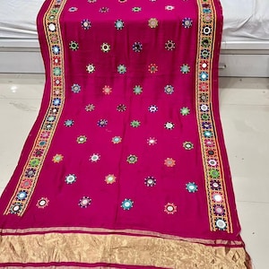 May include: A magenta textile featuring decorative elements. It is embellished with small, circular mirrors and colourful embroidered details. Gold trim borders the edges, adding a touch of elegance. This textile is likely a decorative item, possibly a shawl or scarf.