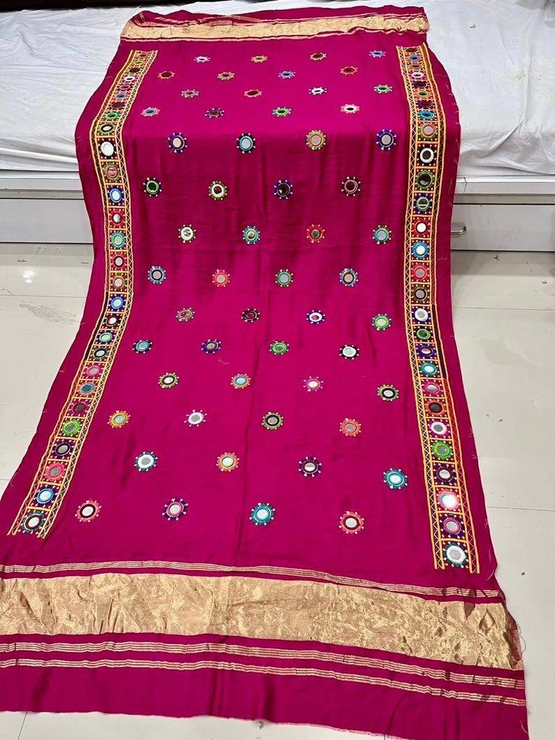 May include: A magenta textile featuring decorative elements. It is embellished with small, circular mirrors and colourful embroidered details. Gold trim borders the edges, adding a touch of elegance. This textile is likely a decorative item, possibly a shawl or scarf.