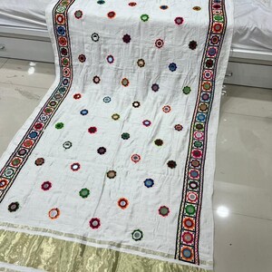 May include: White textile with colourful embroidered floral patterns and mirror accents. The edges feature a detailed border with geometric designs and gold trim. This decorative textile is suitable for home decor.
