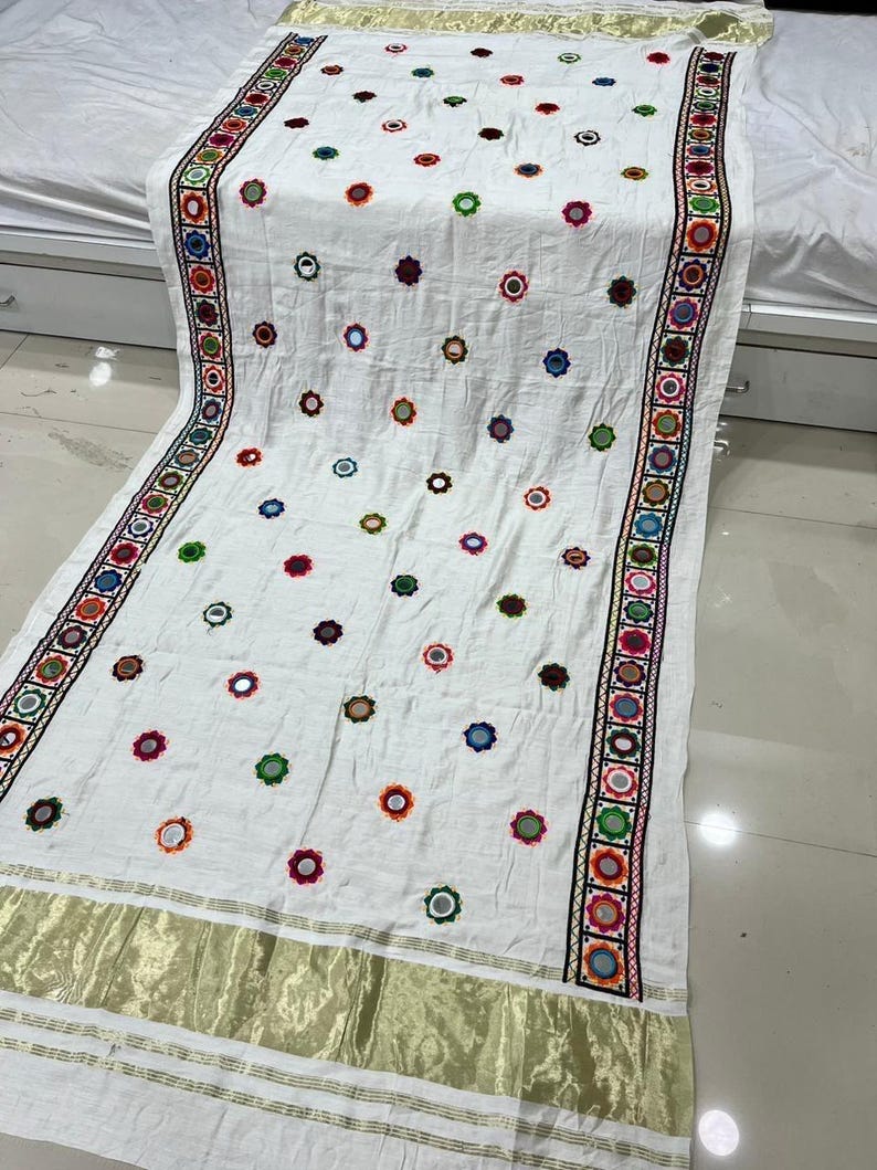 May include: White textile with colourful embroidered floral patterns and mirror accents. The edges feature a detailed border with geometric designs and gold trim. This decorative textile is suitable for home decor.