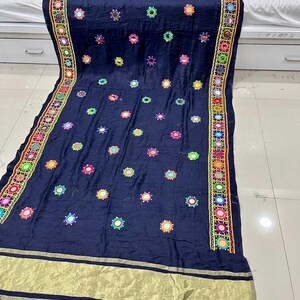 May include: A navy blue textile featuring embroidered floral patterns and small mirrors. The edges have a decorative border in various colours. A gold-coloured band runs along the bottom edge.