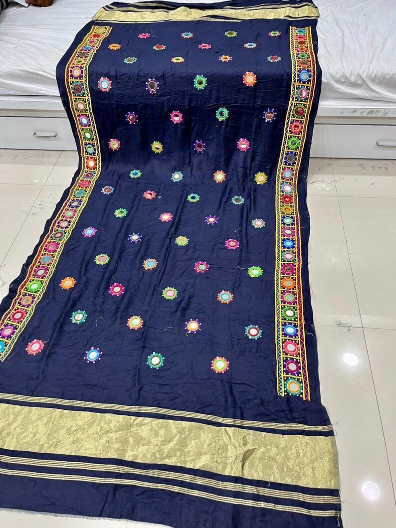 May include: A navy blue textile featuring embroidered floral patterns and small mirrors. The edges have a decorative border in various colours. A gold-coloured band runs along the bottom edge.
