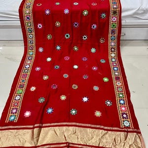 May include: A striking red textile adorned with mirrors and embroidery. The fabric is embellished with small, circular mirrors and colorful embroidery. A gold border with geometric patterns and embroidery frames the edges of the textile.