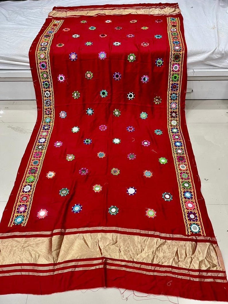May include: A striking red textile adorned with mirrors and embroidery. The fabric is embellished with small, circular mirrors and colorful embroidery. A gold border with geometric patterns and embroidery frames the edges of the textile.