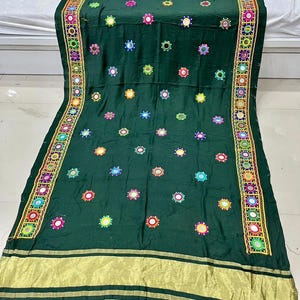 May include: A dark green textile featuring embroidered circular designs in various colours and a gold border. The fabric is decorated with small, round, mirrored accents and a decorative border with intricate patterns. The textile is likely a decorative cloth or garment.