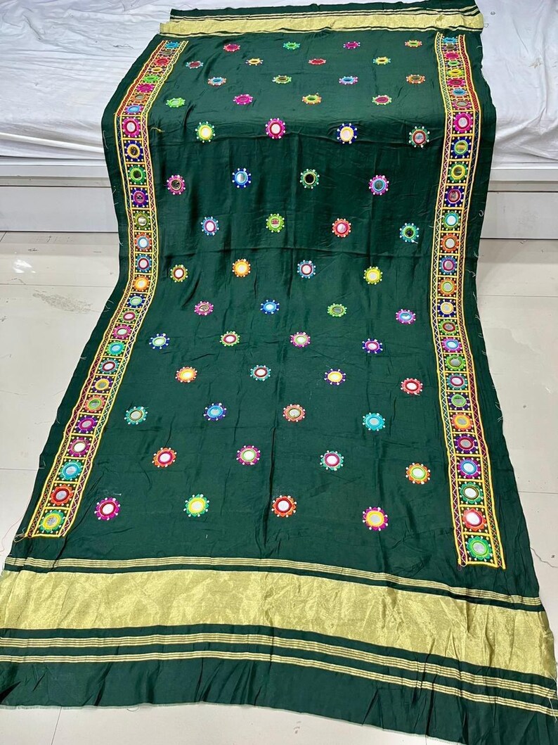 May include: A dark green textile featuring embroidered circular designs in various colours and a gold border. The fabric is decorated with small, round, mirrored accents and a decorative border with intricate patterns. The textile is likely a decorative cloth or garment.