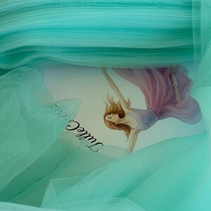 May include: A close-up of a roll of light teal tulle fabric. The fabric is layered, creating a textured effect. A card with a ballerina illustration is partially visible beneath the tulle. The card has the word "Tulle" on it.