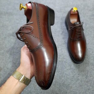 May include: A pair of dark brown leather oxford shoes featuring a polished finish, lace-up closures, and decorative brogue detailing along the seams and toe cap. Each shoe includes a wooden shoe tree insert to maintain its structured shape.