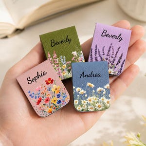 Op de afbeelding: Vier rechthoekige magnetische boekenleggers met bloemenprints en namen. Eén is olijfgroen met 'Beverly', één is lavendelkleurig met 'Beverly', één is roze met 'Sophia' en één is blauw met 'Andrea'. Elk heeft een uniek ontwerp met wilde bloemen.