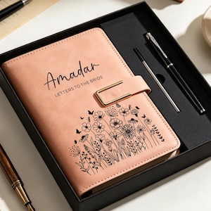 Pode incluir: Um diário cor de pêssego com um fecho de metal dourado, apresentando o texto impresso a preto "Amadar LETTERS TO THE BRIDE" e uma ilustração floral de linhas pretas. O diário é apresentado numa caixa de presente preta com uma caneta preta.