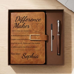 May include: A tan leather-like journal with a gold-tone metal clasp, featuring the printed text Difference Maker and a definition ending with See also: Sophia. The journal is presented in a brown gift box alongside a matching pen and a thin metal stylus.
